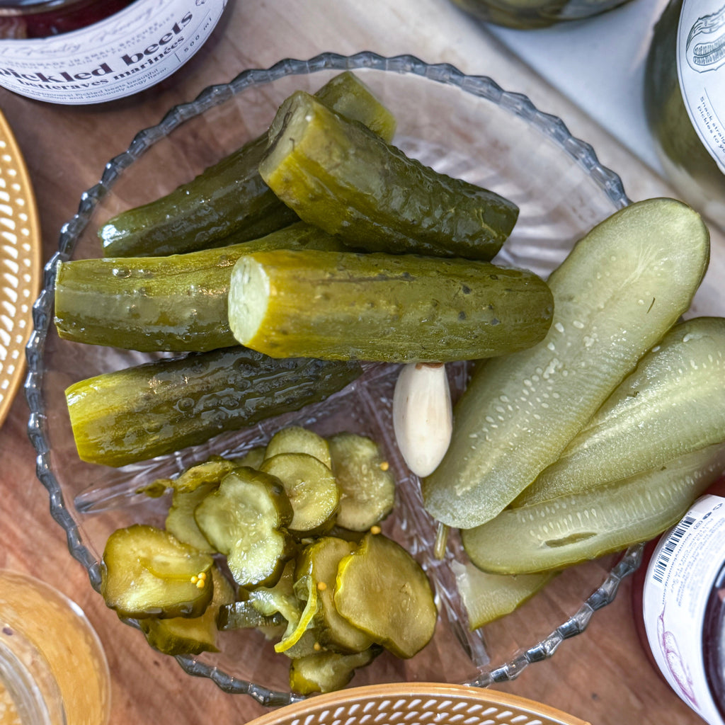 Glass bowl filled with pickles on a wooden surface with jars in the background.