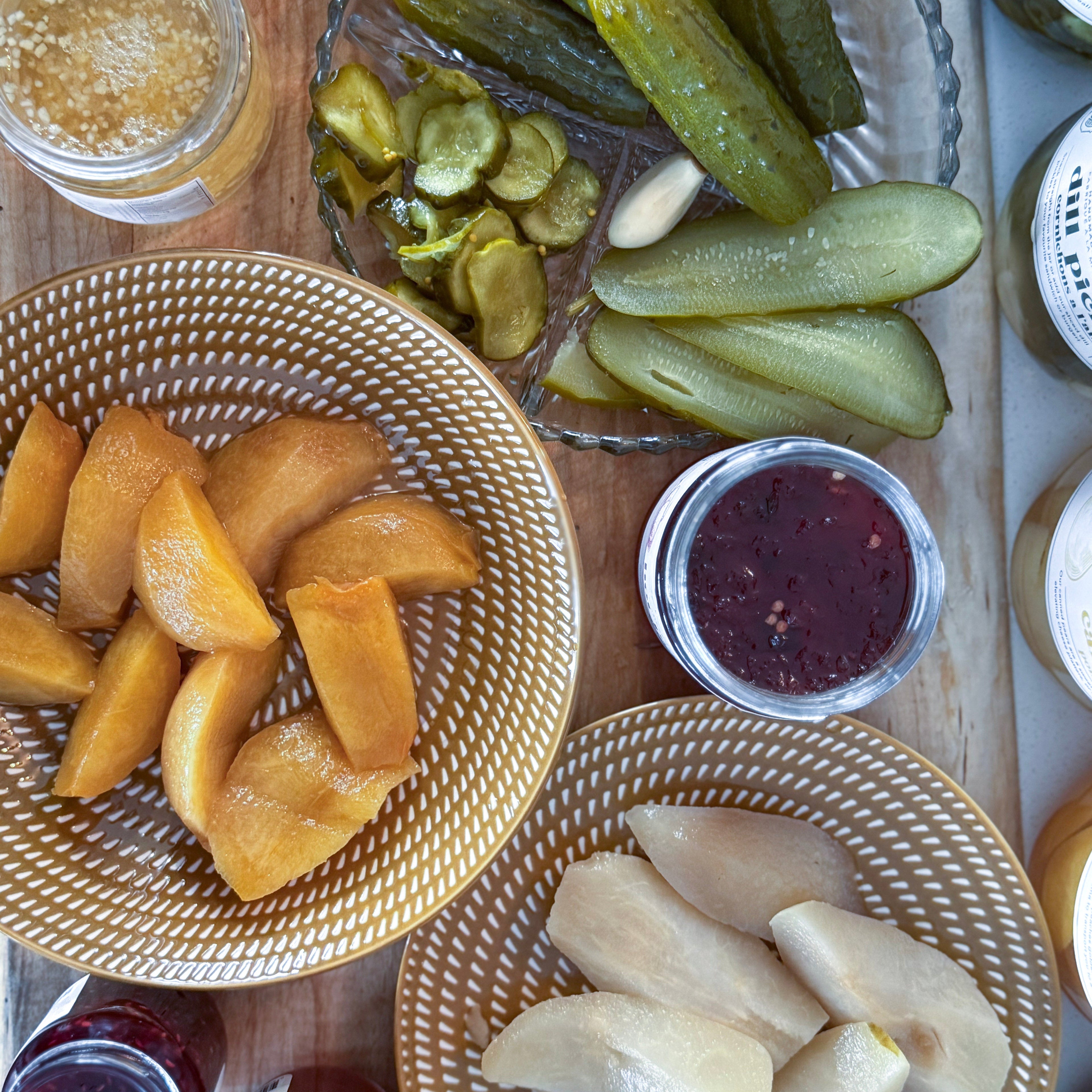 Assorted pickled vegetables and fruits on a wooden board with jars of preserves.