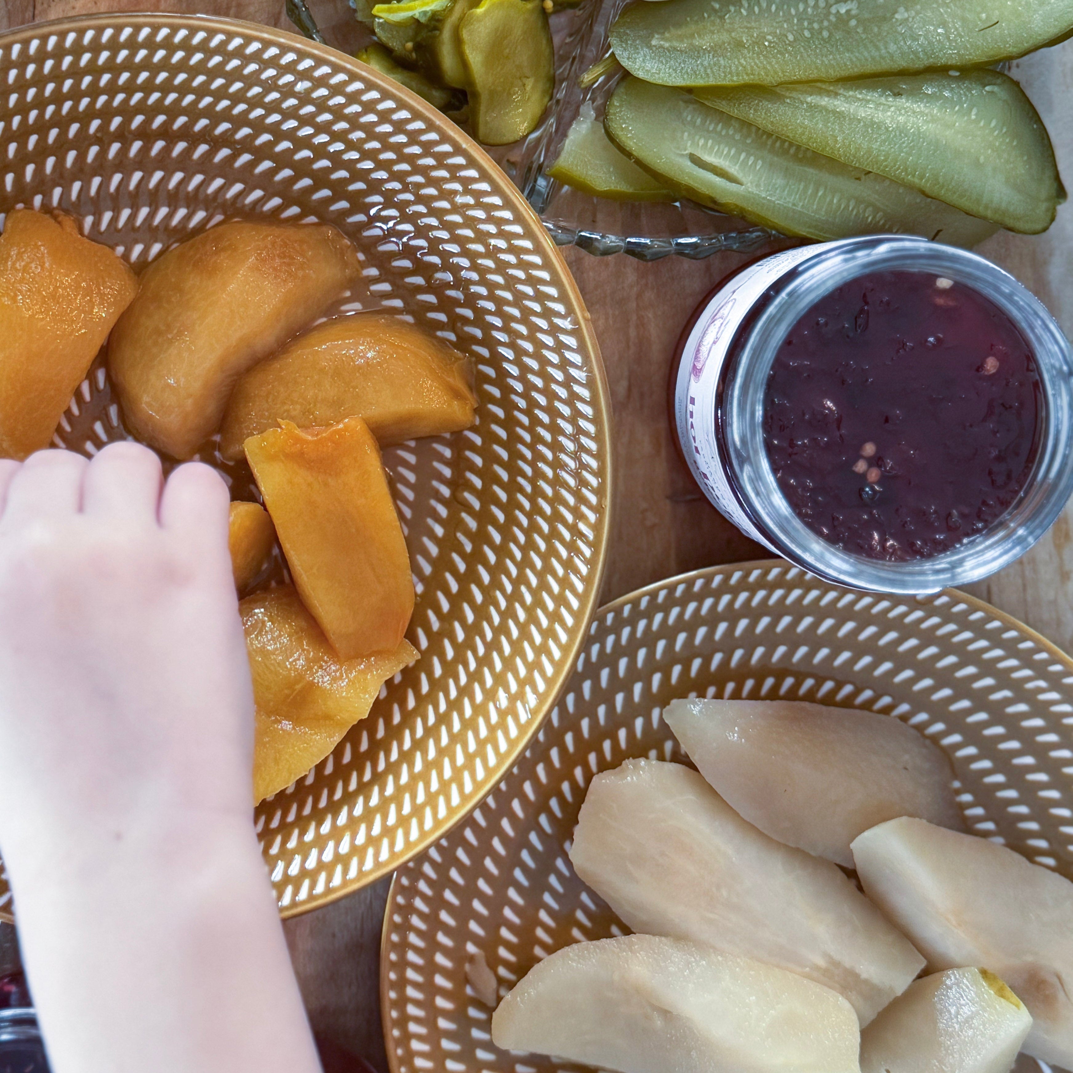 Assorted canned fruit and pickled vegetables on a wooden table with a hand reaching for a peach.