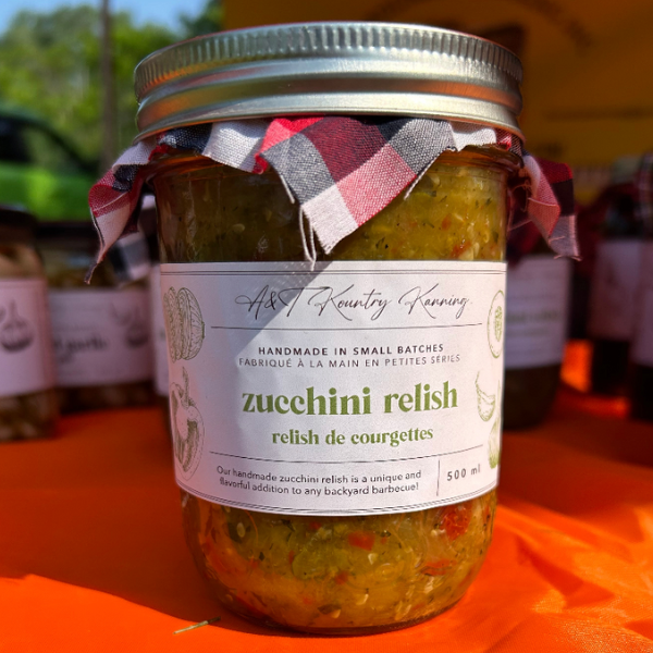 A jar of Zucchini Relish with a red and black checkered cloth on top, set against a table with other jars in the background.